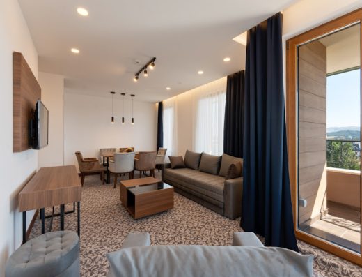 Dnevna soba apartmana u Apart hotel & SPA Queen of Zlatibor sa sivim kaučem, trpezarijskim delom, TV-om na zidu, tamnim zavesama i pogledom na planine kroz balkonska vrata