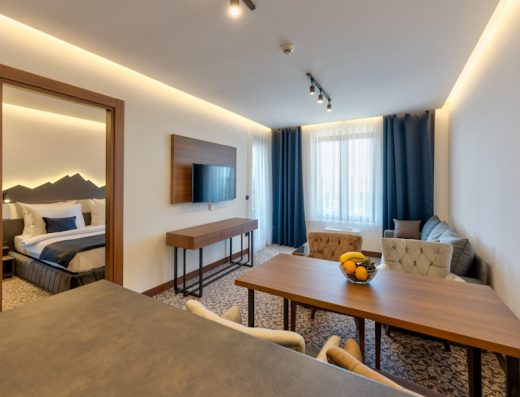 Apartman u Apart hotel & SPA Queen of Zlatibor sa pogledom iz dnevne sobe ka spavaćoj sobi, TV-om, drvenim nameštajem, plavim zavesama i voćem na stolu