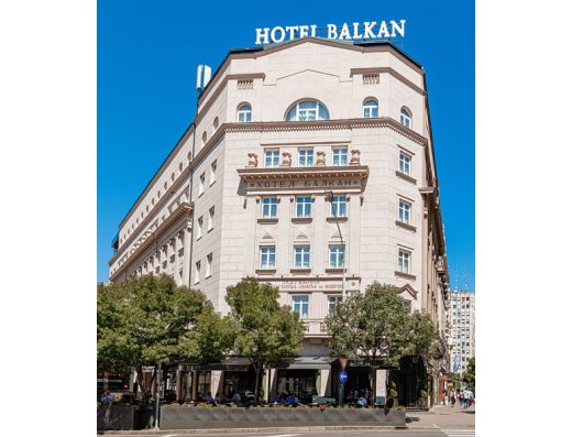 Fasada Hotel Balkan sa natpisom na vrhu zgrade, belom kamenom fasadom, drvoredima i pogledom na ulicu po sunčanom danu