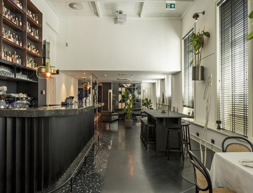 Bar u Hotel Indigo Belgrade sa tamnim terazo pultom, policama sa bocama, mesing lampom, visokim stolicama i visećim biljkama