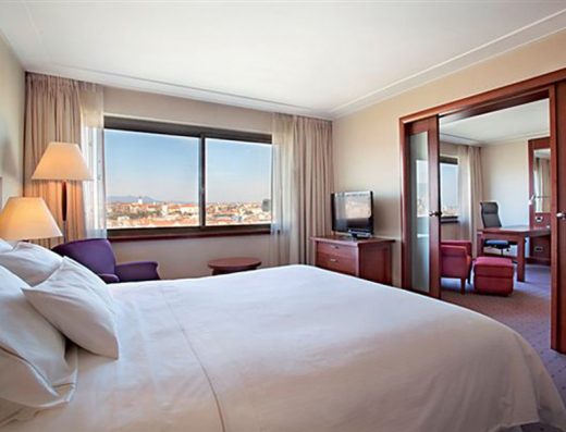 Spavaća soba apartmana u Hotel Westin Zagreb sa bračnim krevetom, belom posteljinom, ljubičastom foteljom, radnim stolom i pogledom na grad