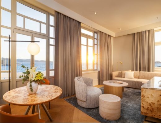 Dnevni boravak apartmana u Hyatt Regency Zadar sa elegantnim nameštajem u bež tonovima, mermernim stolom, cvećem i panoramskim pogledom na more pri zalasku sunca