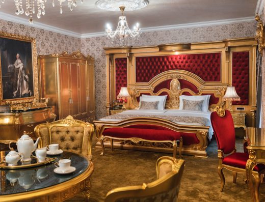Luksuzni apartman u Prezident Palace Belgrade sa velikim crvenim tapaciranim uzglavljem, zlatnim baroknim nameštajem, kristalnim lusterom, zlatnim ormanom i umetničkom slikom