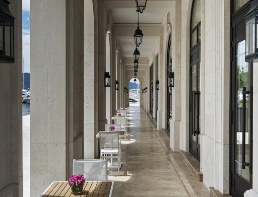 Arkadni hodnik u Regent Porto Montenegro sa kamenim stubovima, crnim lampionima, belim stolovima i stolicama i pogledom na marinu