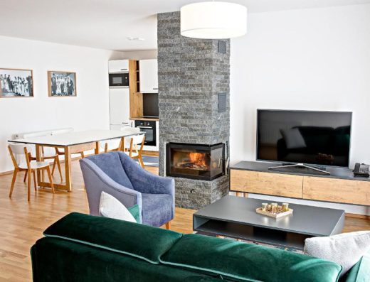 Dnevna soba apartmana u Tarčin Forest Resort & Spa Sarajevo hotelu sa kaminom od kamena, zelenim kaučem, sivom foteljom, TV-om i kuhinjom sa trpezarijom