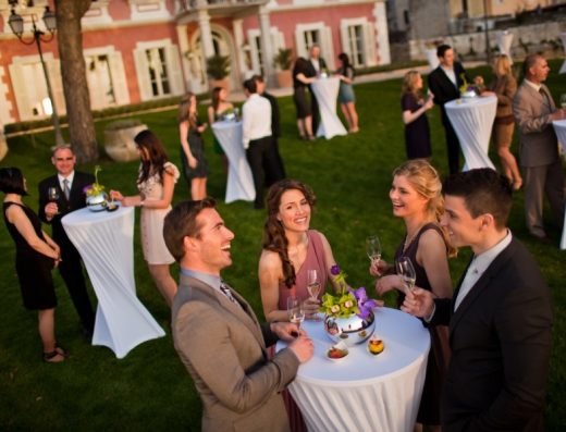 Večernji koktail događaj u vrtu Villa Polesini sa gostima, visokim koktail stolovima, šampanjcem i roze fasadom vile u pozadini